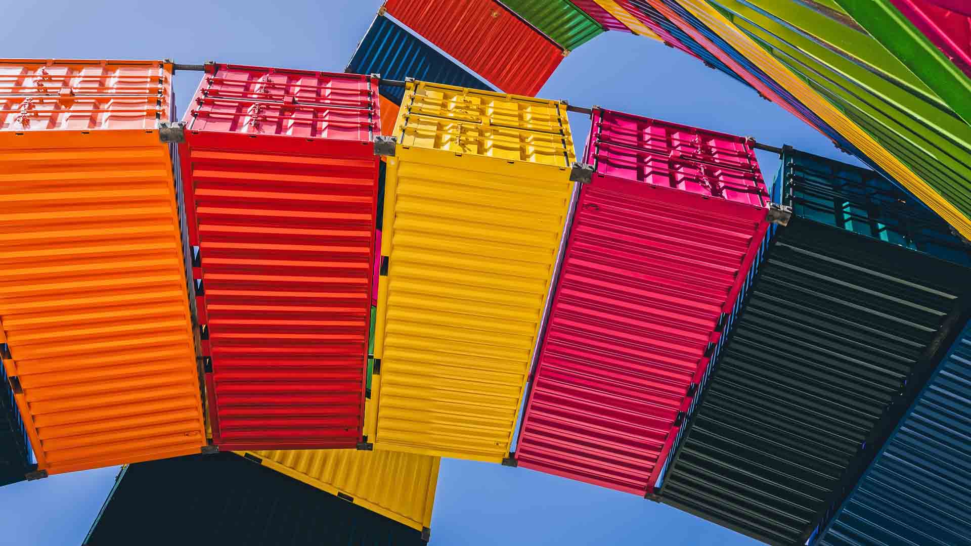 Contêineres de transporte coloridos, que lembram artigos vibrantes, estão dispostos em um arco contra um céu azul claro.