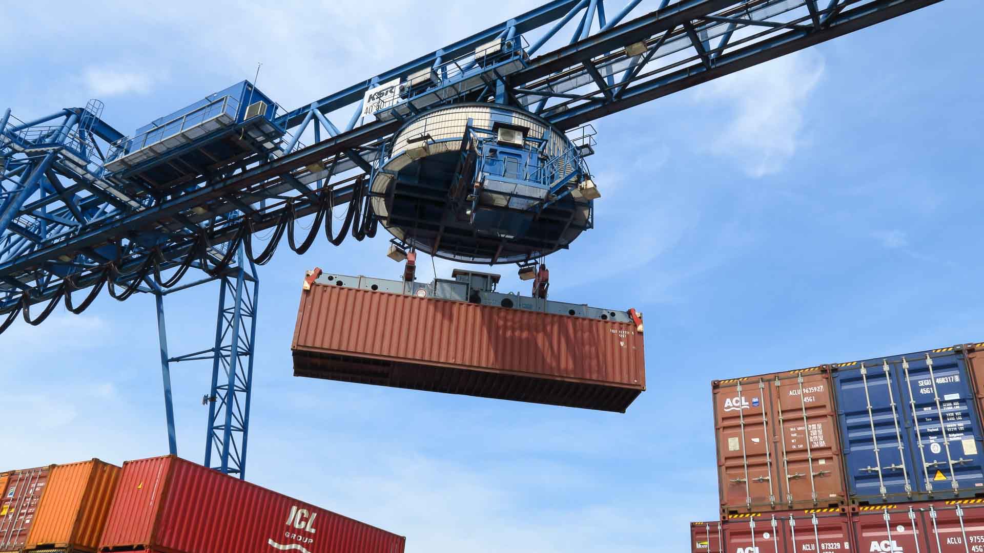 Em um porto movimentado, um guindaste iça um grande contêiner vermelho, provavelmente cheio de "Aviamentos da China", bem acima do local. Abaixo, outros contêineres sugerem a dança movimentada de importação de aviamentos sob um céu azul claro.