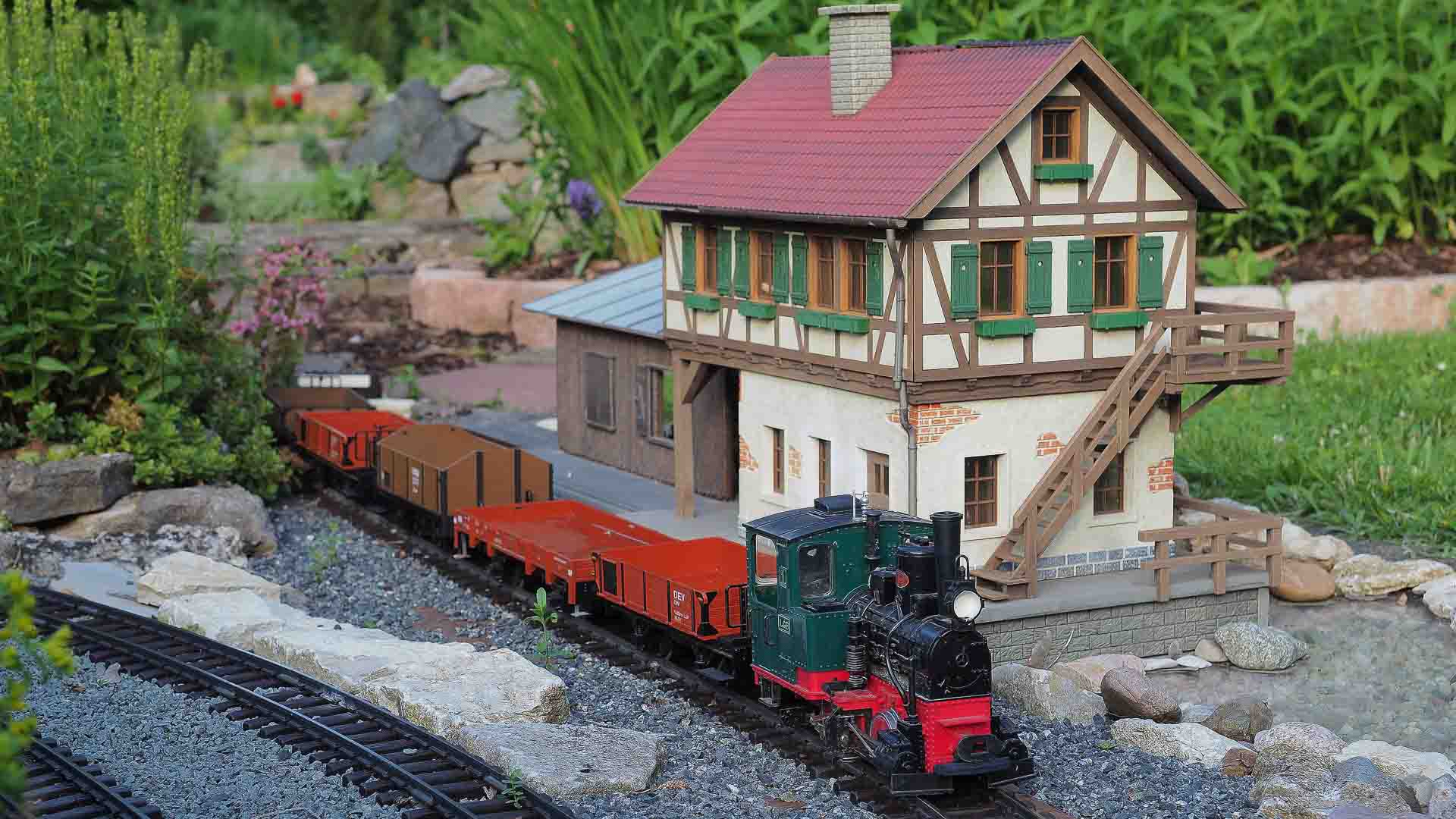 Um modelo de trem com locomotiva a vapor e vagões de carga, transportando itens de importação em miniatura, passa por um pequeno prédio de modelos cercado por vegetação e pedras.