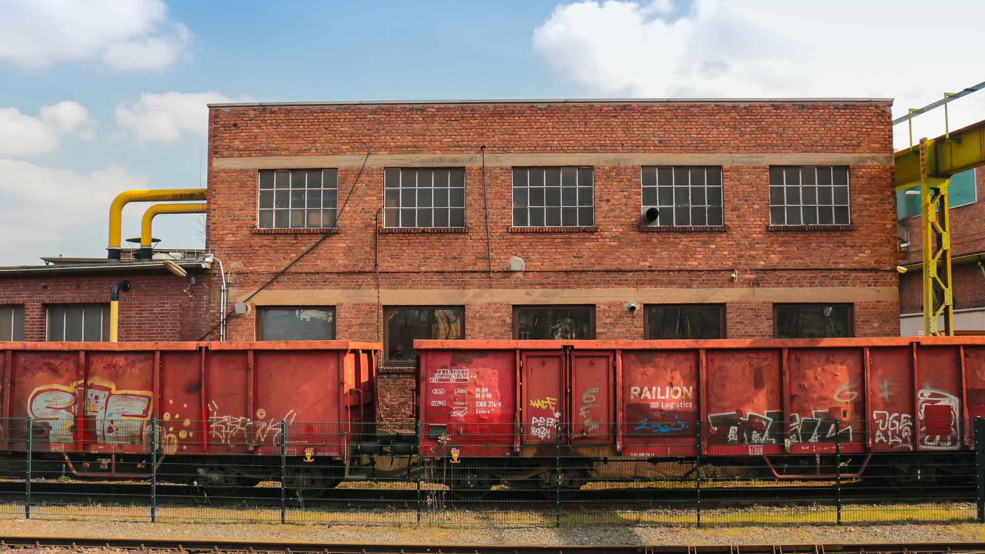 Em um dia ensolarado, um prédio industrial de tijolos com grandes janelas se ergue atrás de dois vagões de trem de carga cobertos de grafite vermelho, lembrando guias sobre como fazer arte de rua ou o mundo colorido da importação de brinquedos.