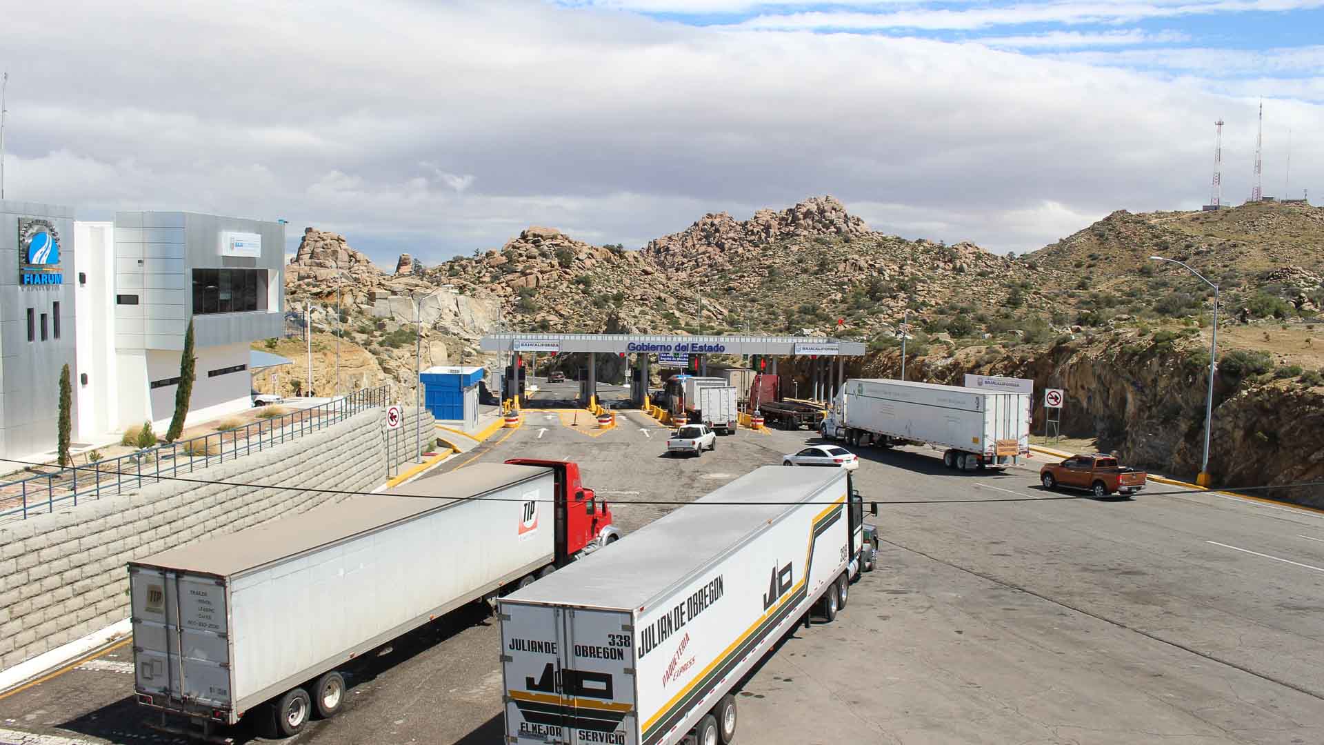 Caminhões se aproximam de um posto de controle de fronteira em uma área montanhosa sob um céu nublado, possivelmente transportando equipamentos acadêmicos por essa rota de importação vital.