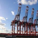 Quatro grandes guindastes vermelhos estão no porto sob um céu azul com nuvens dispersas, símbolos de importação movimentada em 2025. Um pequeno barco flutua em primeiro plano, sugerindo o fluxo contínuo da Equipamentos de Tecnologia por essas águas movimentadas.