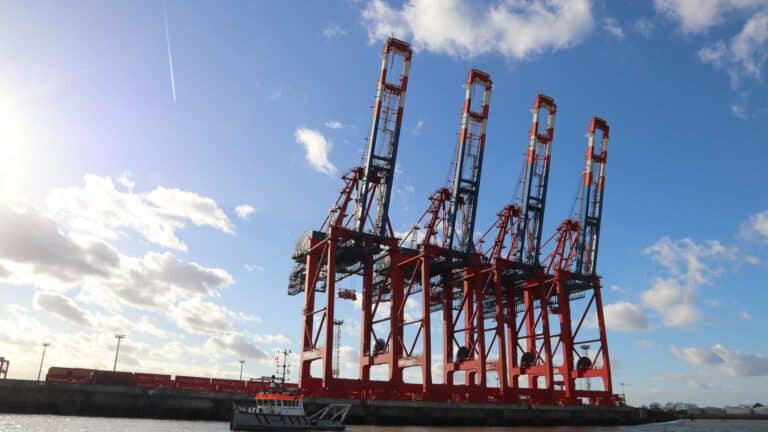Quatro grandes guindastes vermelhos estão no porto sob um céu azul com nuvens dispersas, símbolos de importação movimentada em 2025. Um pequeno barco flutua em primeiro plano, sugerindo o fluxo contínuo da Equipamentos de Tecnologia por essas águas movimentadas.