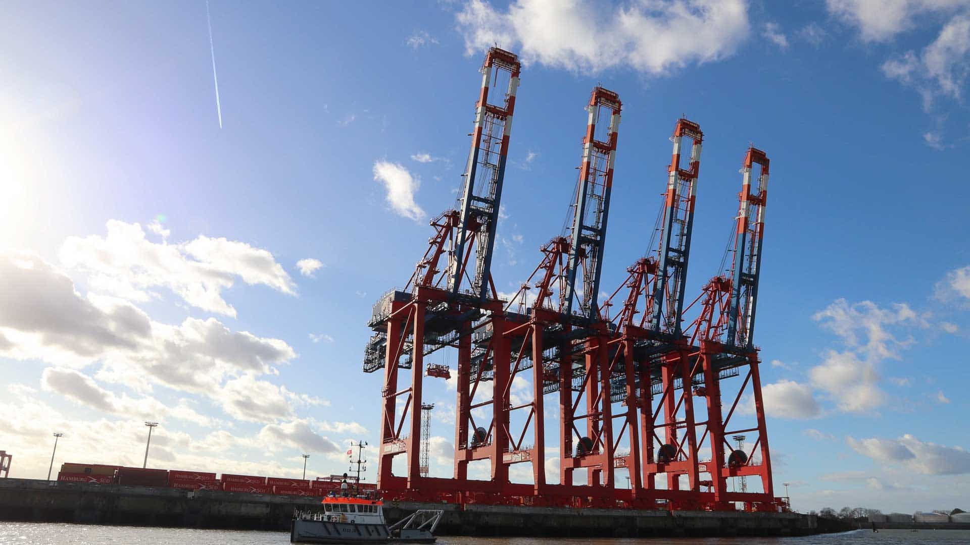 Quatro grandes guindastes vermelhos estão no porto sob um céu azul com nuvens dispersas, símbolos de importação movimentada em 2025. Um pequeno barco flutua em primeiro plano, sugerindo o fluxo contínuo da Equipamentos de Tecnologia por essas águas movimentadas.