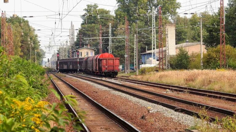 Um trem de carga com vagões vermelhos e marrons, possivelmente cheios de produtos importados da China, viaja sobre trilhos ferroviários por uma área rural adornada com vegetação e pontilhada por linhas de energia.