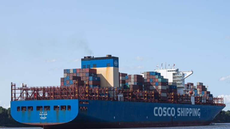 Um grande navio porta-contêineres com "COSCO SHIPPING" na lateral, transportando vários contêineres de carga empilhados, incluindo aqueles para importação de jogos, está se movendo em um corpo d'água.
