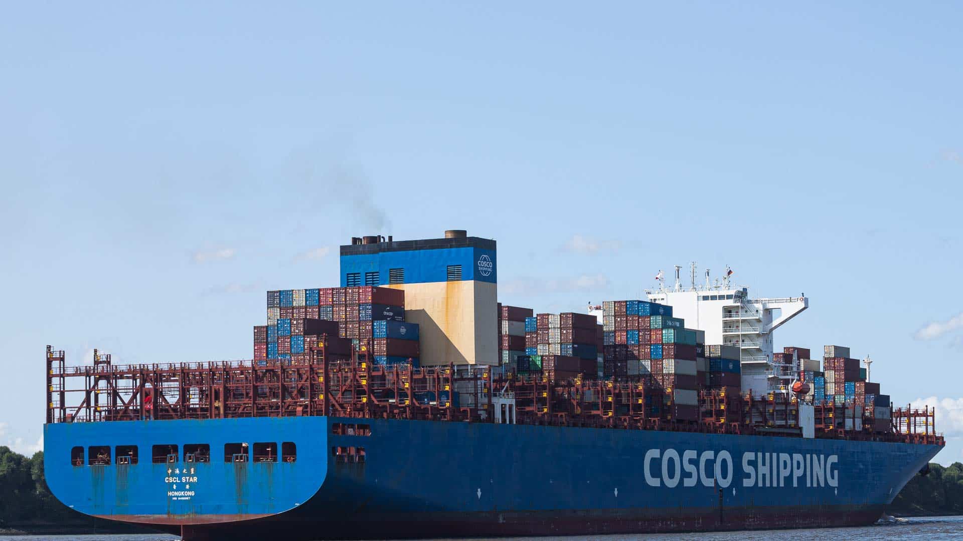 Um grande navio porta-contêineres com "COSCO SHIPPING" na lateral, transportando vários contêineres de carga empilhados, incluindo aqueles para importação de jogos, está se movendo em um corpo d'água.