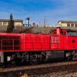 Uma locomotiva a diesel vermelha repousa orgulhosamente sobre os trilhos da ferrovia, enquanto edifícios altos emolduram a cena contra um céu azul claro, lembrando possibilidades futuras, como hoverboards voando acima.