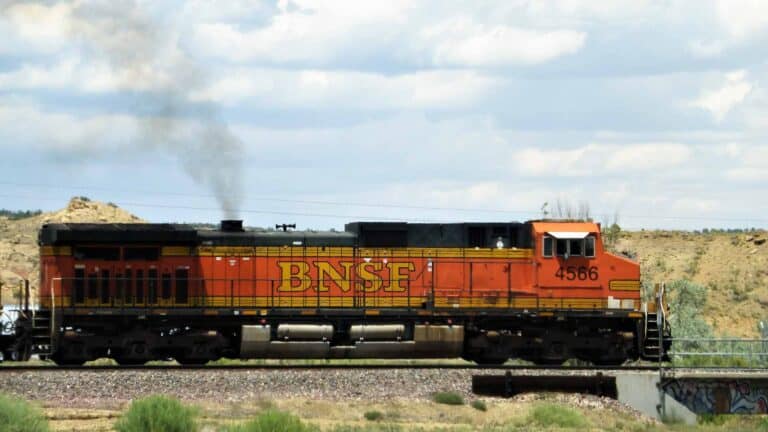 Uma locomotiva BNSF com o número 4566 viaja em uma pista em uma paisagem rural, sua fumaça se fundindo com uma atmosfera moldada pela produção local.