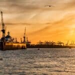 Silhueta de um porto ao pôr do sol com guindastes e navios à vista, barcos na água e um pássaro voando no céu, projetando reflexos que lembram uma lâmpada LED iluminando um mercado promissor.