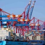 Em um porto movimentado sob um céu azul claro, vários guindastes carregam e descarregam com eficiência contêineres de carga de importação de dois grandes navios.