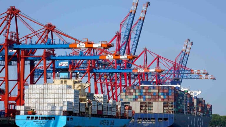 Em um porto movimentado sob um céu azul claro, vários guindastes carregam e descarregam com eficiência contêineres de carga de importação de dois grandes navios.