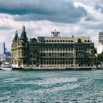 Vista da histórica Estação de Trem Haydarpaşa em Istambul, Turquia, do outro lado do mar, com um céu nublado e uma paisagem urbana ao fundo — uma cena que poderia facilmente preencher páginas de cadernos de viagem com dicas essenciais para qualquer aventureiro que busca capturar seu encanto atemporal.