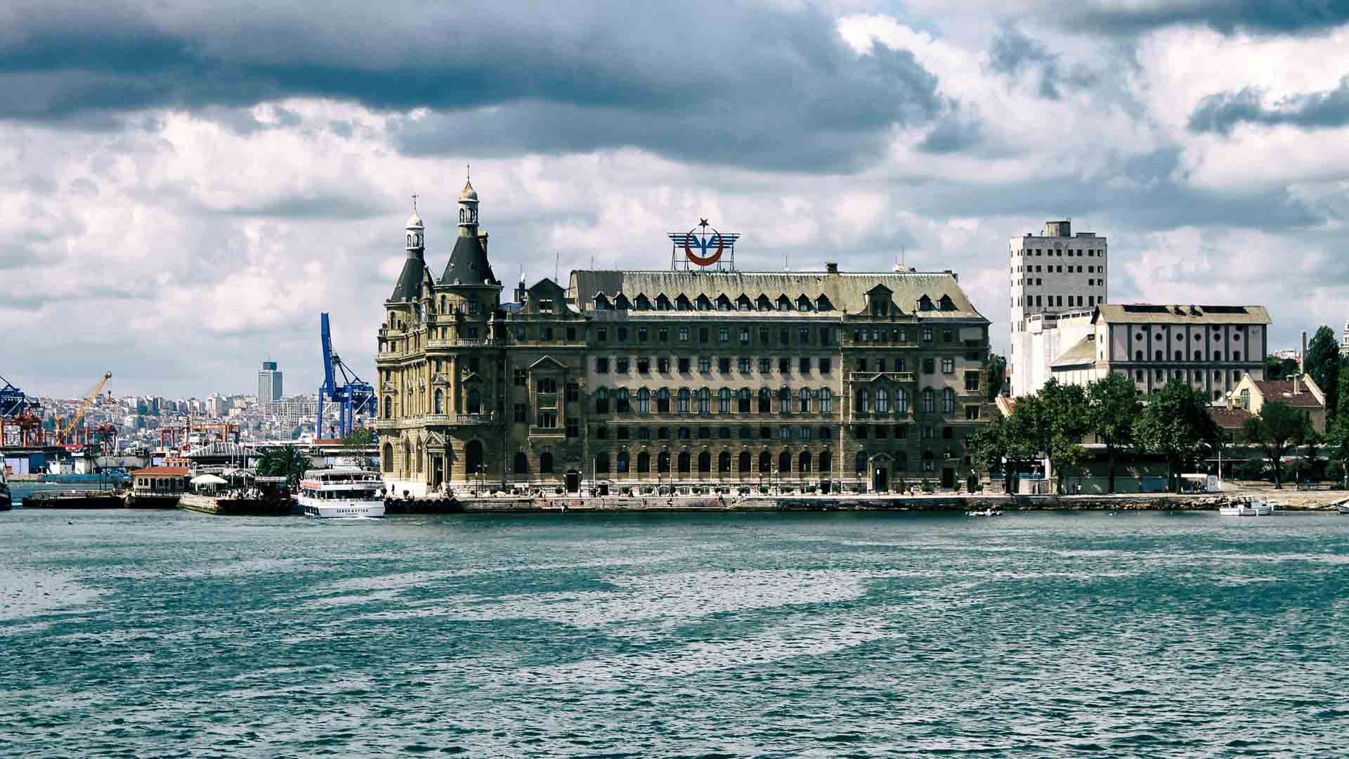 Vista da histórica Estação de Trem Haydarpaşa em Istambul, Turquia, do outro lado do mar, com um céu nublado e uma paisagem urbana ao fundo — uma cena que poderia facilmente preencher páginas de cadernos de viagem com dicas essenciais para qualquer aventureiro que busca capturar seu encanto atemporal.