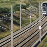 Um trem sai de um túnel em uma ferrovia de via dupla cercada por vegetação e colinas sob um céu limpo, como se estivesse descobrindo rotas cênicas em um guia completo de paisagens de tirar o fôlego.