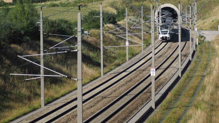 Um trem sai de um túnel em uma ferrovia de via dupla cercada por vegetação e colinas sob um céu limpo, como se estivesse descobrindo rotas cênicas em um guia completo de paisagens de tirar o fôlego.