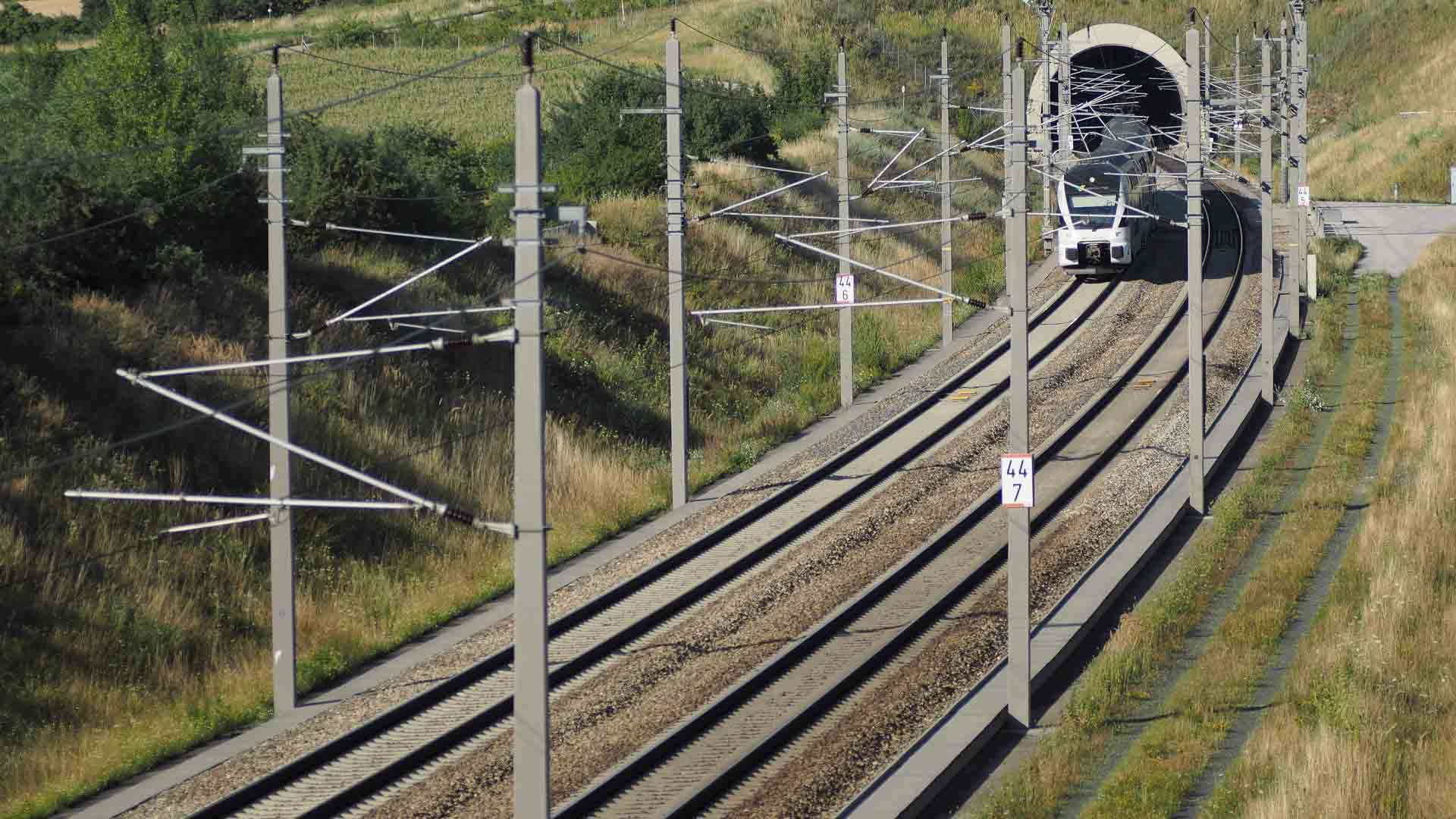 Um trem sai de um túnel em uma ferrovia de via dupla cercada por vegetação e colinas sob um céu limpo, como se estivesse descobrindo rotas cênicas em um guia completo de paisagens de tirar o fôlego.