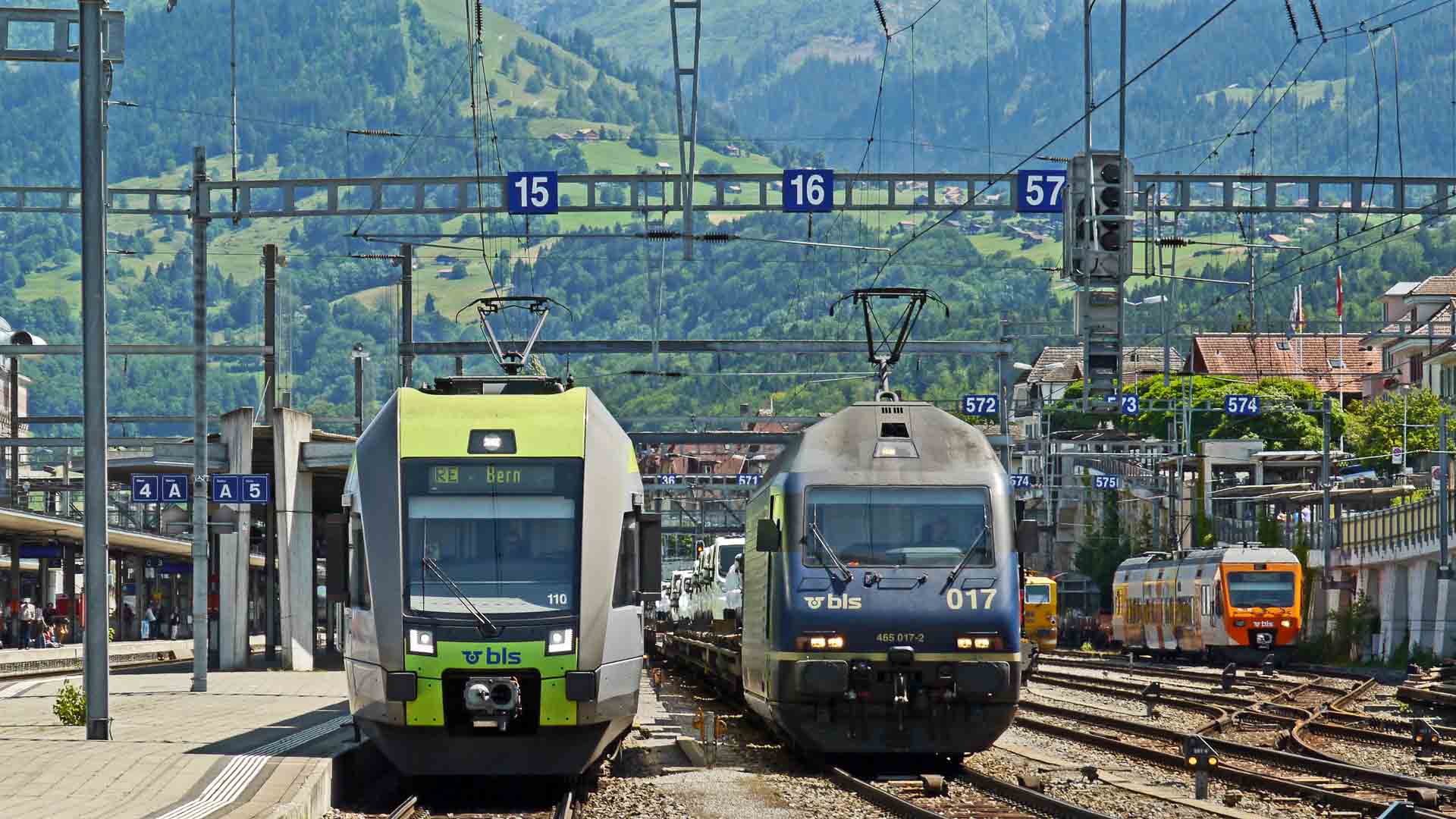 Dois trens em uma estação ferroviária com fios aéreos, cercados por montanhas e uma pequena cidade ao fundo, são alimentados parcialmente por painéis solares. Este toque moderno oferece dicas sobre viagens sustentáveis e itens essenciais de importação para viajantes ecologicamente conscientes.