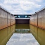 Uma eclusa de canal com grandes paredes de concreto está cheia de água turva, refletindo a estrutura. O céu acima está nublado. Em meio a esse cenário industrial, oportunidades inexploradas para energia renovável, como a importação de painéis solares, surgem, prometendo um futuro mais brilhante.