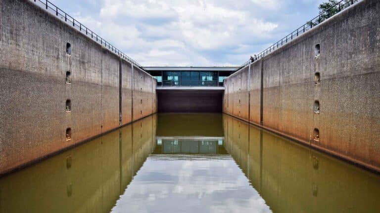 Uma eclusa de canal com grandes paredes de concreto está cheia de água turva, refletindo a estrutura. O céu acima está nublado. Em meio a esse cenário industrial, oportunidades inexploradas para energia renovável, como a importação de painéis solares, surgem, prometendo um futuro mais brilhante.