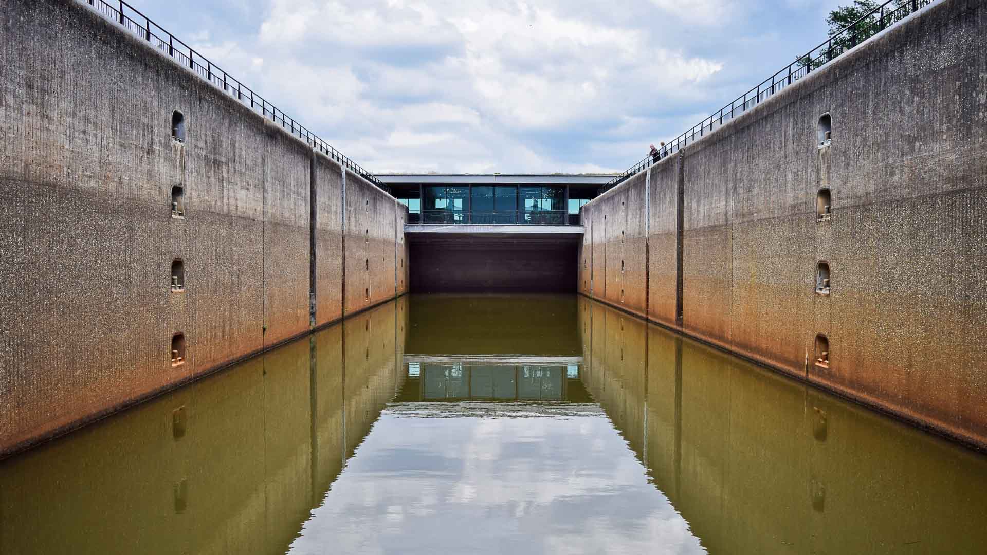 Uma eclusa de canal com grandes paredes de concreto está cheia de água turva, refletindo a estrutura. O céu acima está nublado. Em meio a esse cenário industrial, oportunidades inexploradas para energia renovável, como a importação de painéis solares, surgem, prometendo um futuro mais brilhante.