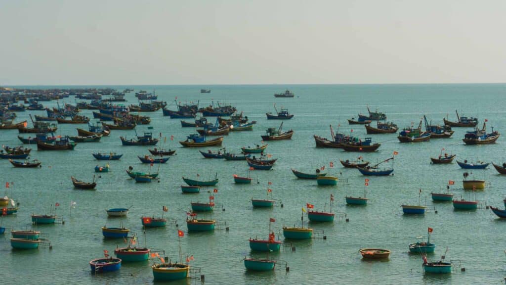 Vários barcos de pesca com bandeiras estão ancorados em águas calmas e azul-turquesa sob um céu nublado, criando um cenário saído de uma coleção Papéis de Parede de 2024.