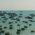 Vários barcos de pesca com bandeiras estão ancorados em águas calmas e azul-turquesa sob um céu nublado, criando um cenário saído de uma coleção Papéis de Parede de 2024.