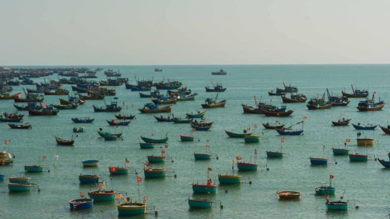 Vários barcos de pesca com bandeiras estão ancorados em águas calmas e azul-turquesa sob um céu nublado, criando um cenário saído de uma coleção Papéis de Parede de 2024.