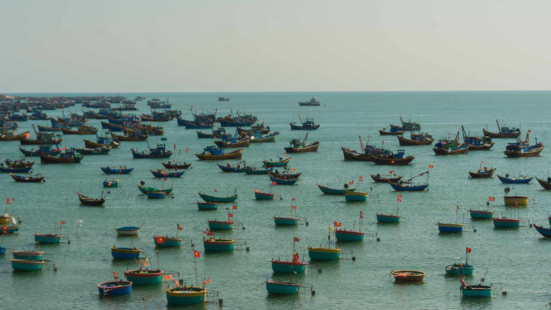 Vários barcos de pesca com bandeiras estão ancorados em águas calmas e azul-turquesa sob um céu nublado, criando um cenário saído de uma coleção Papéis de Parede de 2024.