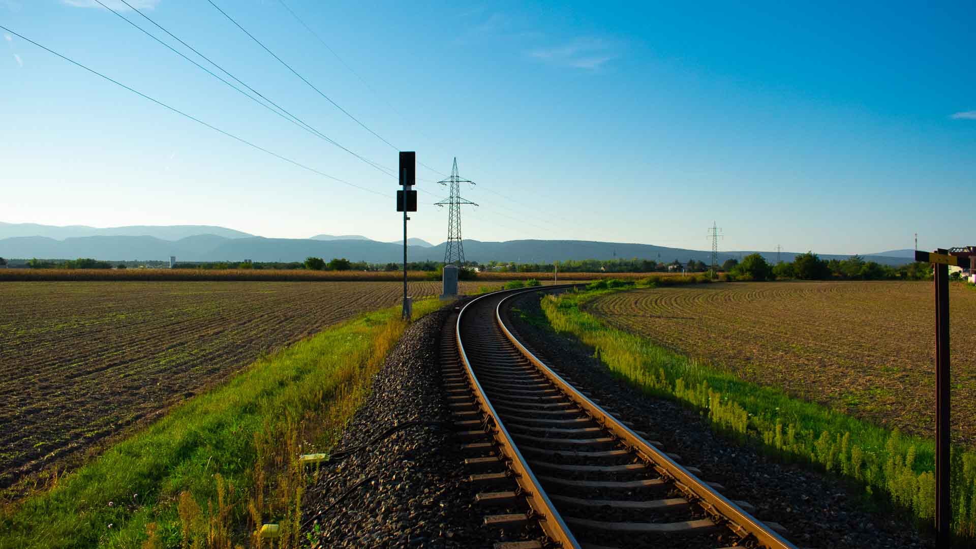 Uma linha ferroviária curva serpenteia por uma paisagem rural, pontilhada por campos e montanhas ao longe, sob um céu azul claro, enquanto scooters passam ocasionalmente, adicionando um toque de modernidade a esse cenário sereno.