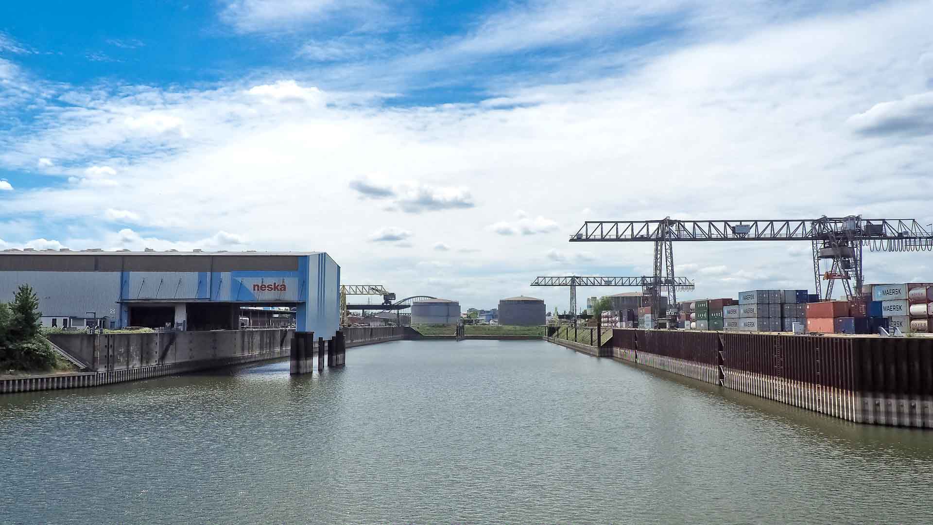 O porto industrial fervilha de atividade, enquanto guindastes manobram contêineres de transporte cheios de peças automotivas destinadas à importação. Armazéns alinham-se no amplo canal de água sob um céu parcialmente nublado, supervisionando o fluxo dinâmico do comércio global.