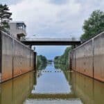 Uma eclusa de canal com águas calmas se estende em direção a uma ponte. Árvores alinham as laterais, e uma estrutura de concreto é visível à esquerda, lembrando rotas de ciclismo no exterior. Para entusiastas, explorar tais rotas fornece Dicas Essenciais para descobrir caminhos cênicos e encontrar joias escondidas Importação.