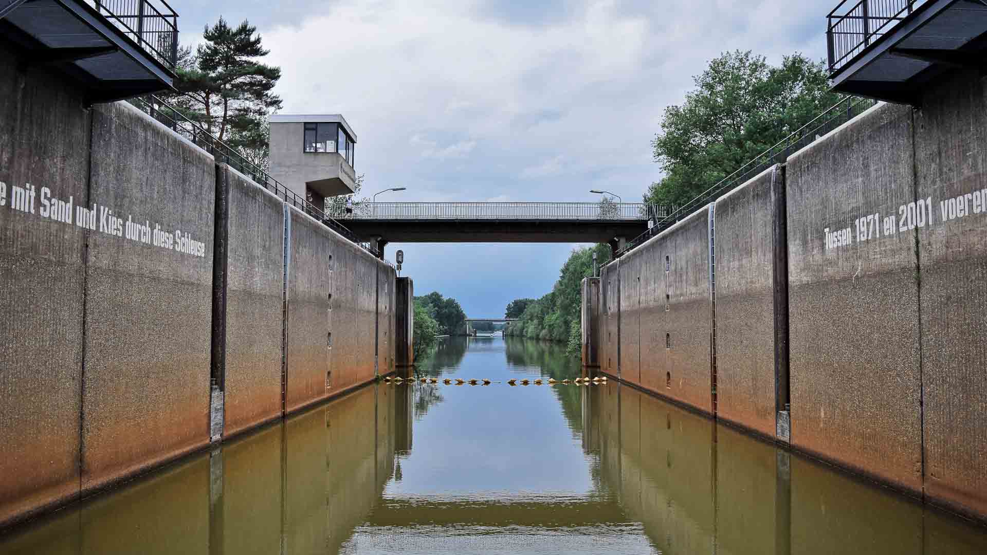 Uma eclusa de canal com águas calmas se estende em direção a uma ponte. Árvores alinham as laterais, e uma estrutura de concreto é visível à esquerda, lembrando rotas de ciclismo no exterior. Para entusiastas, explorar tais rotas fornece Dicas Essenciais para descobrir caminhos cênicos e encontrar joias escondidas Importação.