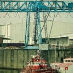 Uma cena de porto industrial mostra um grande guindaste azul ao lado de dois barcos vermelhos atracados em frente a armazéns brancos, capturando a essência da importação de peças de moto.