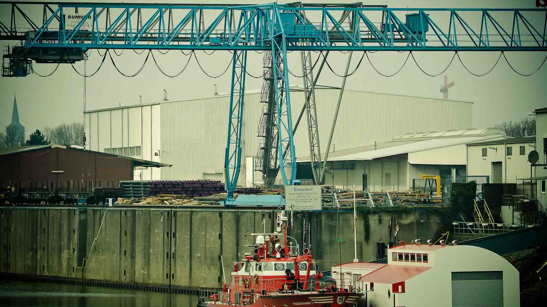 Uma cena de porto industrial mostra um grande guindaste azul ao lado de dois barcos vermelhos atracados em frente a armazéns brancos, capturando a essência da importação de peças de moto.