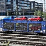 Um trem transportando duas camadas de vagões azuis e vermelhos em um vagão aberto desliza suavemente pelo pátio ferroviário, lembrando um processo simplificado, muito parecido com a importação de perfumes exclusivos. Um edifício se destaca no cenário.