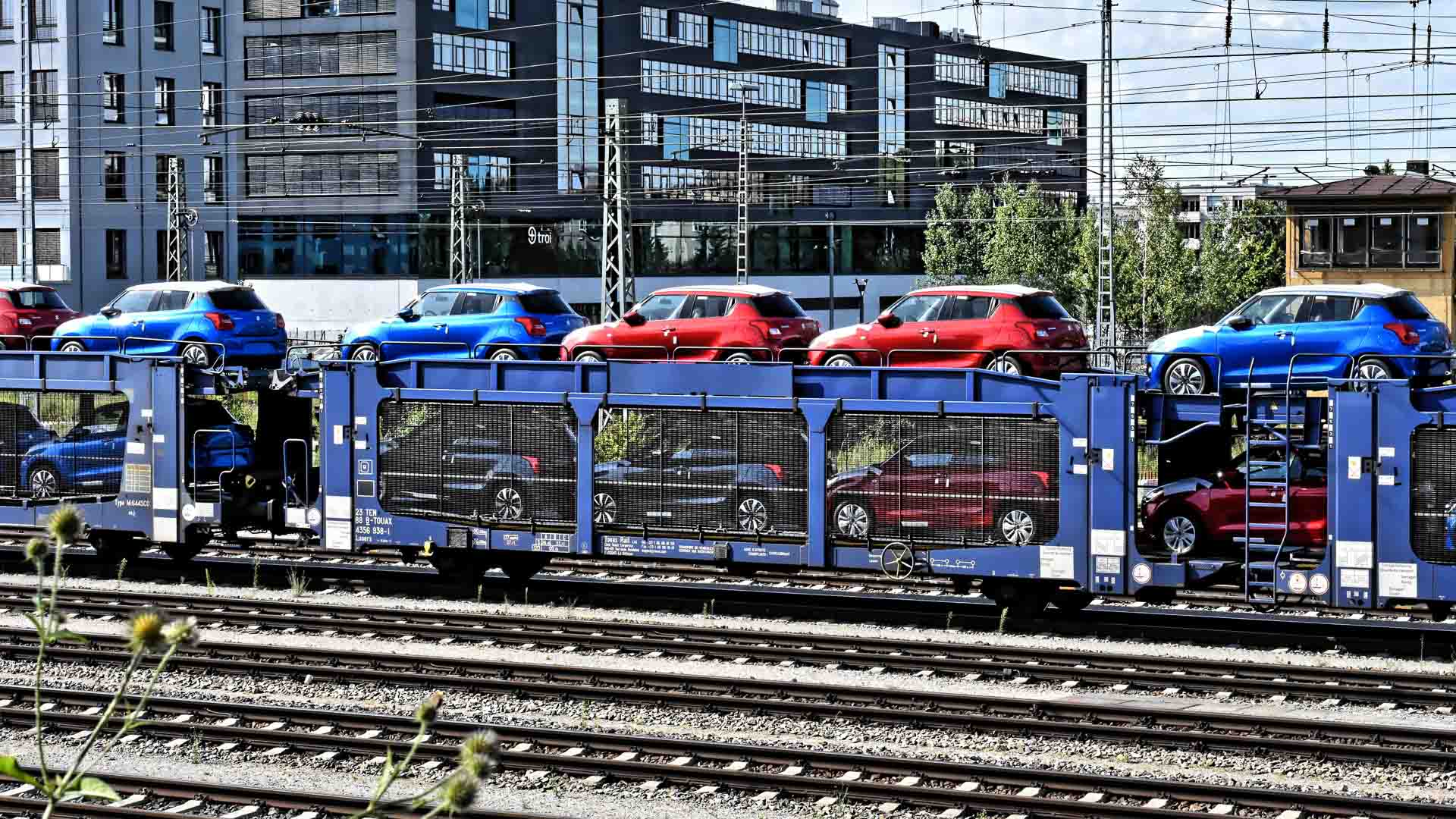 Um trem transportando duas camadas de vagões azuis e vermelhos em um vagão aberto desliza suavemente pelo pátio ferroviário, lembrando um processo simplificado, muito parecido com a importação de perfumes exclusivos. Um edifício se destaca no cenário.