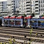 Um trem transportando carros vermelhos e azuis em vagões de plataforma viaja sobre trilhos ferroviários, exibindo produtos prontos para a Importação de Produtos. Prédios de apartamentos modernos são visíveis ao fundo, destacando uma cidade que prospera com inovações certificadas pela Anvisa.