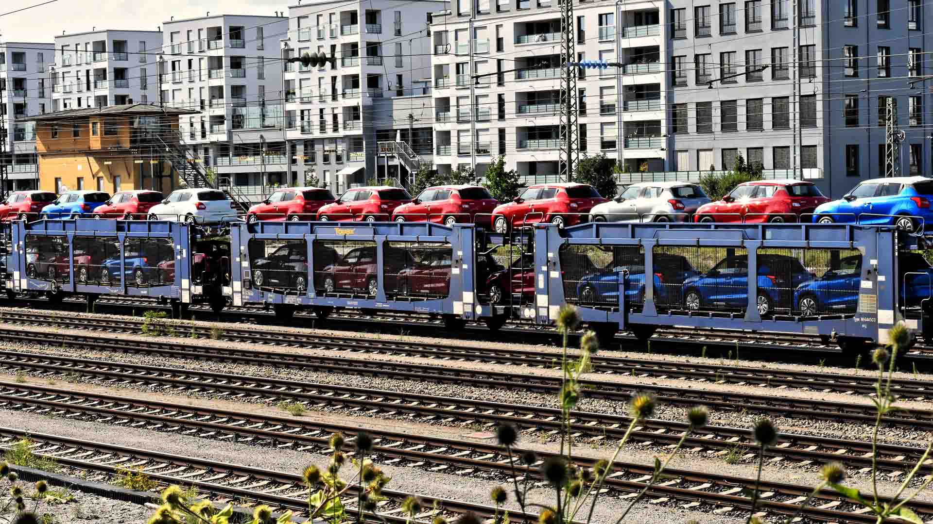 Um trem transportando carros vermelhos e azuis em vagões de plataforma viaja sobre trilhos ferroviários, exibindo produtos prontos para a Importação de Produtos. Prédios de apartamentos modernos são visíveis ao fundo, destacando uma cidade que prospera com inovações certificadas pela Anvisa.