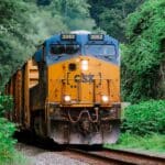 Um trem de carga CSX azul e amarelo, simbolizando produtos inovadores, viaja em uma linha cercada por vegetação verdejante e terreno rochoso, preparando o cenário para uma jornada de importação tranquila até 2025.