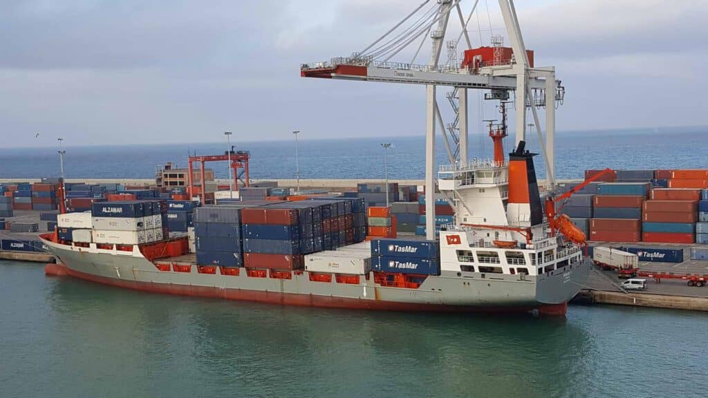 Um navio de carga carregado com contêineres está atracado em um porto sob um grande guindaste. A área do porto, movimentada com atividade de importação, está cheia de contêineres de transporte empilhados, exibindo oportunidades para empresas que buscam expandir seu alcance. Ao fundo, o mar se estende infinitamente.