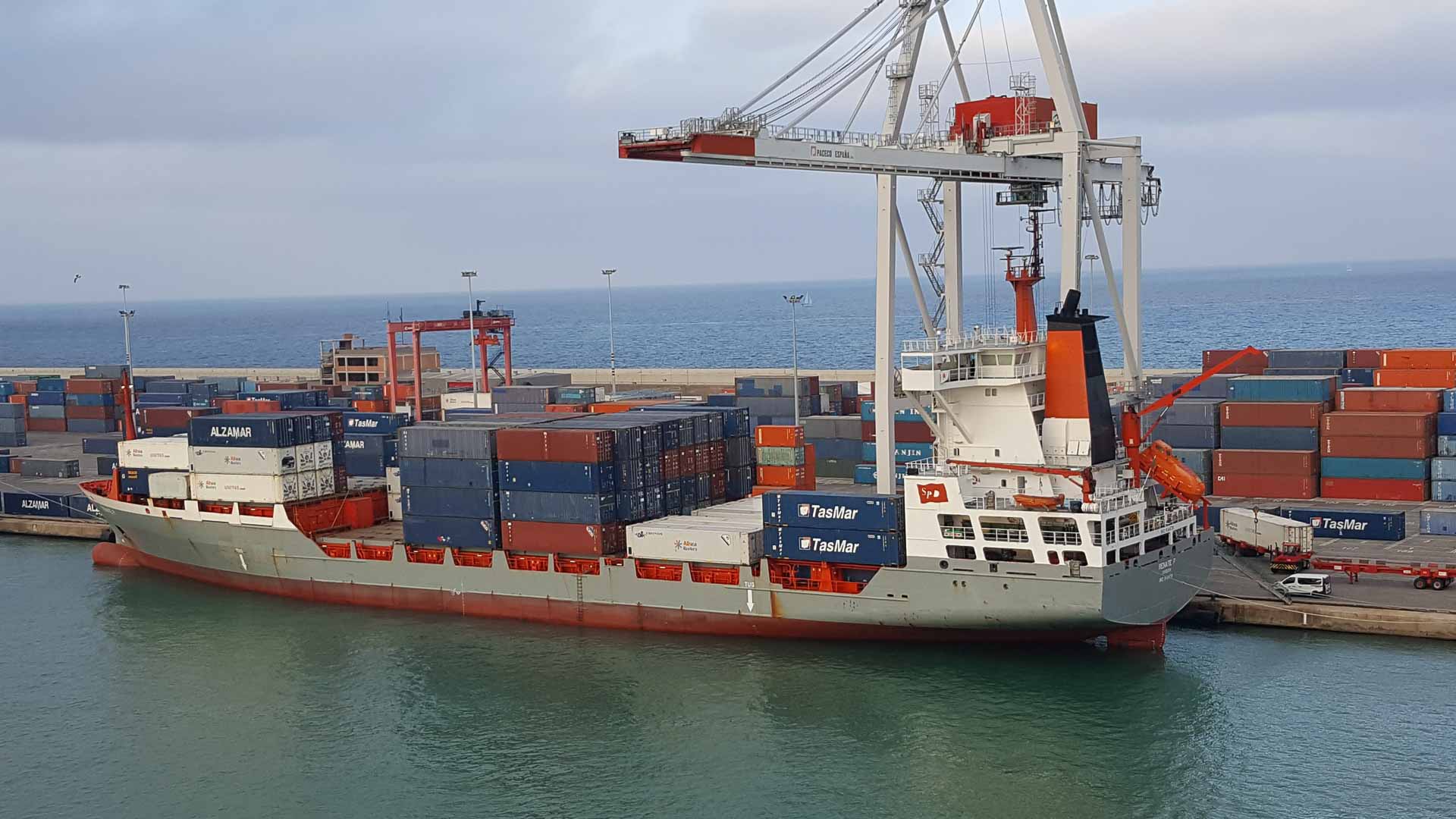Um navio de carga carregado com contêineres está atracado em um porto sob um grande guindaste. A área do porto, movimentada com atividade de importação, está cheia de contêineres de transporte empilhados, exibindo oportunidades para empresas que buscam expandir seu alcance. Ao fundo, o mar se estende infinitamente.