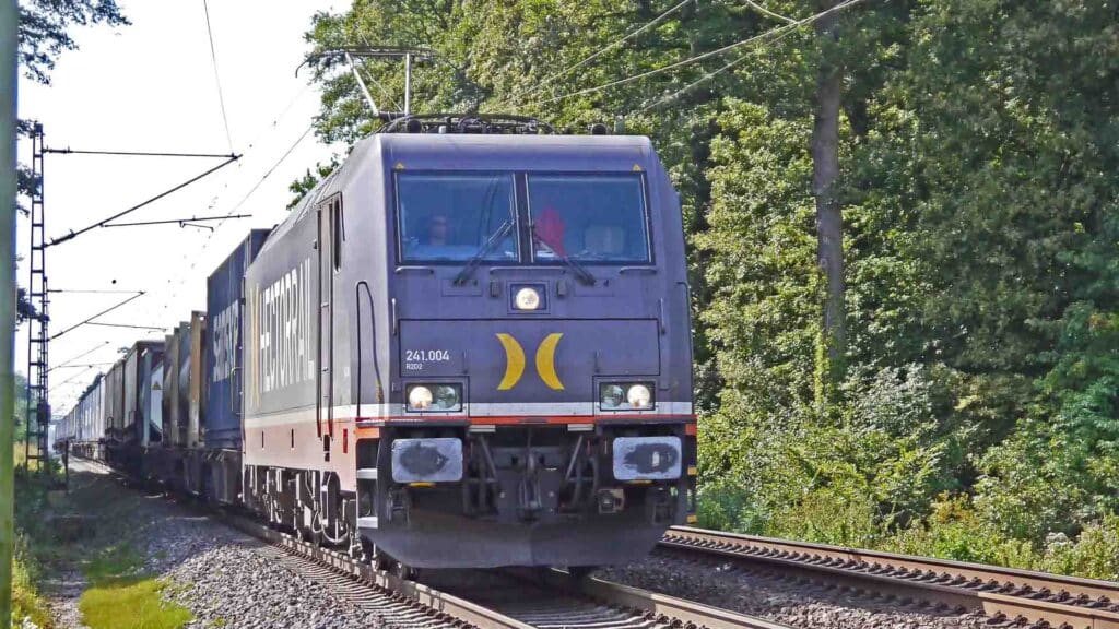 Um trem de carga preto se move por trilhos cercados por vegetação, sob fios elétricos aéreos em um dia ensolarado, simbolizando o futuro da "Importação de Produtos Verdes", conforme descrito no "Guia para 2025".