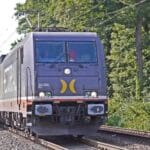 Um trem de carga preto se move por trilhos cercados por vegetação, sob fios elétricos aéreos em um dia ensolarado, simbolizando o futuro da "Importação de Produtos Verdes", conforme descrito no "Guia para 2025".