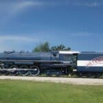 Vista lateral de uma locomotiva a vapor azul e branca, rotulada "Frisco" e "Meteor 4500", exibindo a aplicação da tecnologia ferroviária clássica, sobre trilhos contra um fundo de céu azul claro e vegetação exuberante.