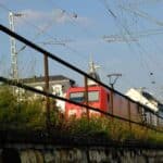 Um trem vermelho se move ao longo dos trilhos ao lado de uma cerca e vegetação alta, evocando as movimentadas rotas de importação de roupas. Prédios e linhas de energia são visíveis ao fundo sob um céu limpo, lembrando as artérias de comércio global.