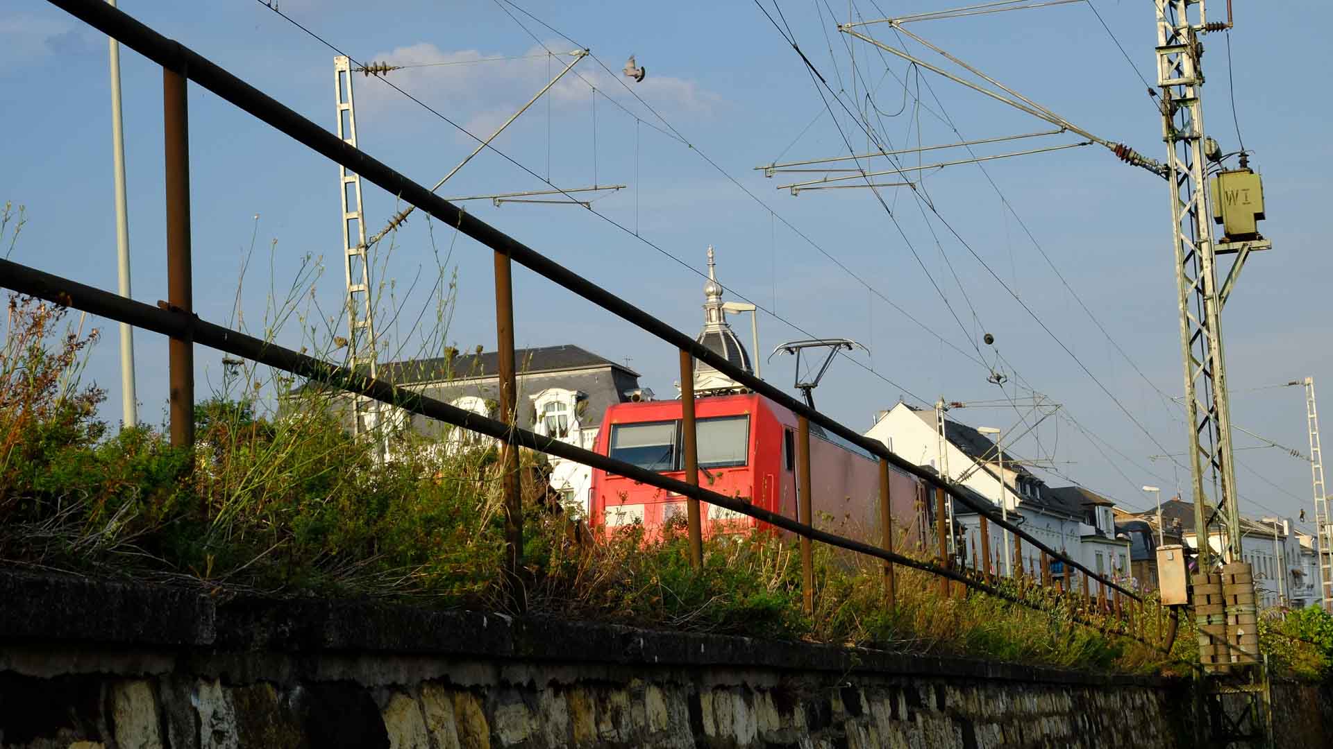 Um trem vermelho se move ao longo dos trilhos ao lado de uma cerca e vegetação alta, evocando as movimentadas rotas de importação de roupas. Prédios e linhas de energia são visíveis ao fundo sob um céu limpo, lembrando as artérias de comércio global.