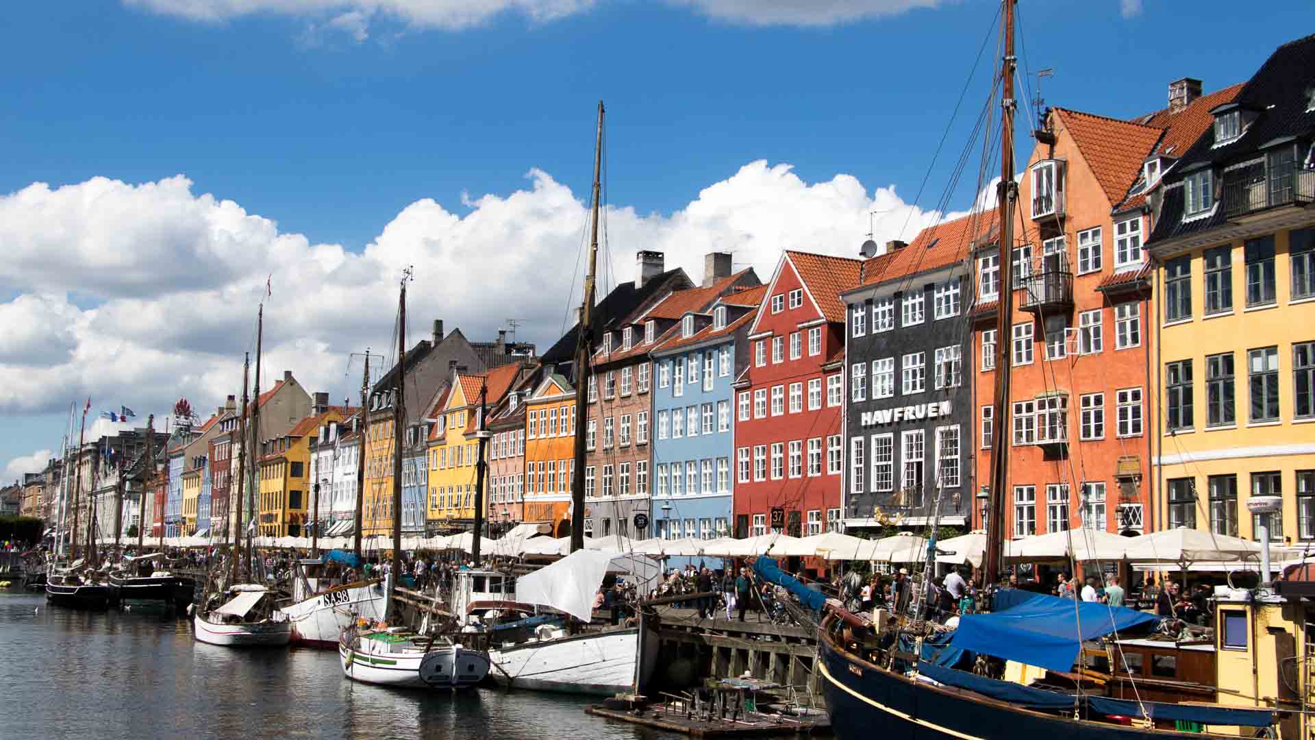 Prédios coloridos e barcos atracados alinham-se em um canal sob um céu parcialmente nublado, evocando o charme da importação. O cenário vibrante é adornado com tecidos e acessórios que refletem a rica tapeçaria cultural da área.