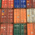 Contêineres de transporte empilhados em várias cores, incluindo vermelho, laranja, azul e branco, com logotipos e números de identificação visíveis. Muitos desses contêineres são parte do processo de importação da China.