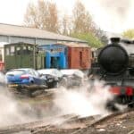 A locomotiva a vapor libera uma nuvem de vapor nos trilhos, enquanto vários carros estacionados e vagões de trem ficam ao fundo. Sob um céu nublado com árvores visíveis, evoca nostalgia em meio às tendências modernas de sustentabilidade.